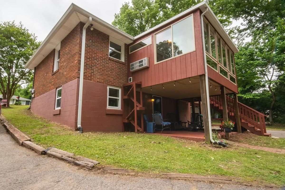 The lower level apartment features a private entrance with a covered deck overlooking the green forest. Large windows allow natural light into the space. A gentle sloping pathway leads to the entrance, and seating areas are visible on the deck.