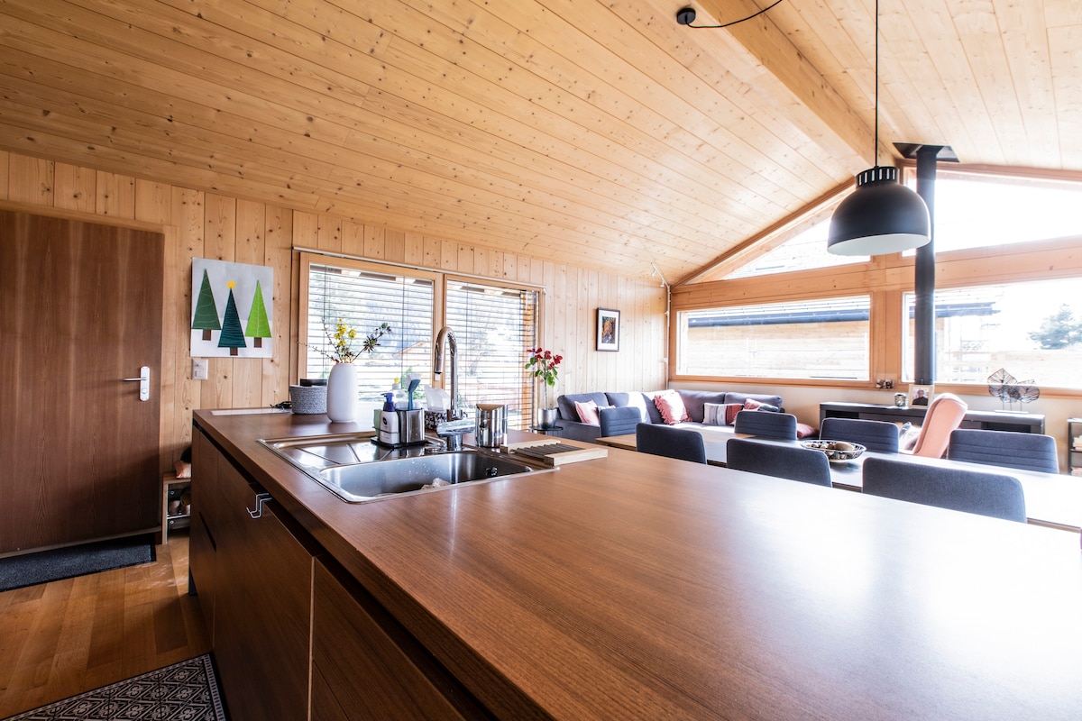 An open kitchen area features a wooden ceiling and a large countertop with a sink. Sunlight enters through large windows, illuminating a cozy seating arrangement with neutral-toned furniture. Decorative elements, including potted plants and artwork, enhance the inviting environment.