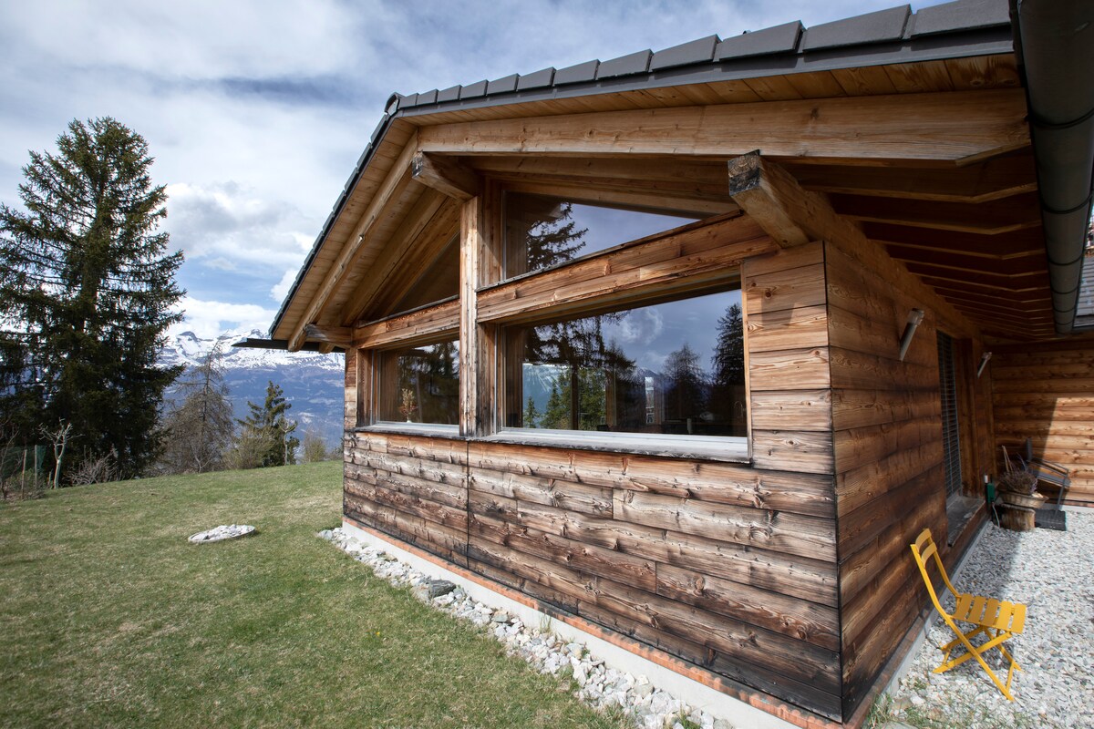 The exterior of a wooden chalet showcases a spacious design with large windows offering expansive views. A well-maintained lawn surrounds the building, complemented by a gravel pathway. A single yellow chair is positioned on the terrace, inviting relaxation.