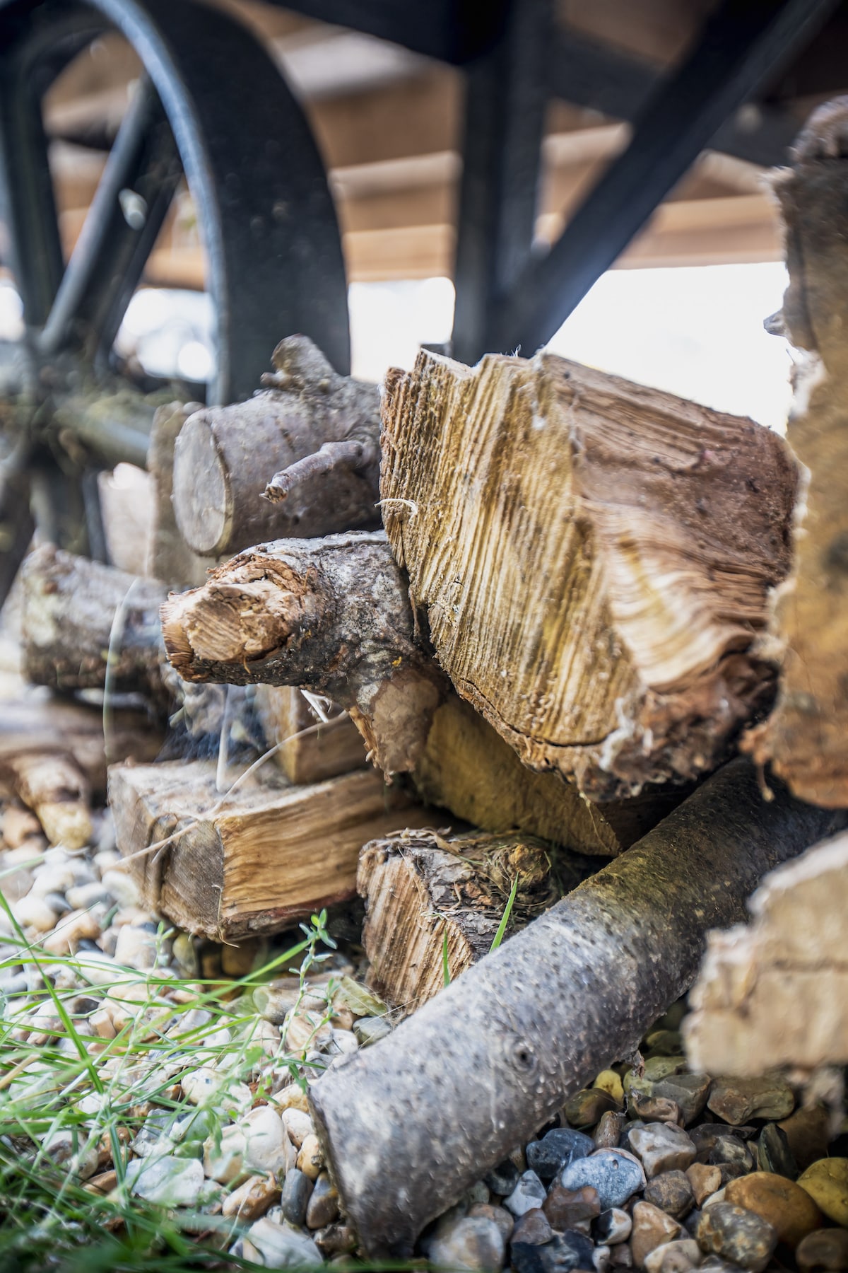 A stack of logs is arranged near a wheel of a wooden structure. The logs vary in size and texture, resting on a bed of smooth pebbles and patches of grass. The arrangement suggests a cozy, rustic element, inviting warmth and comfort.