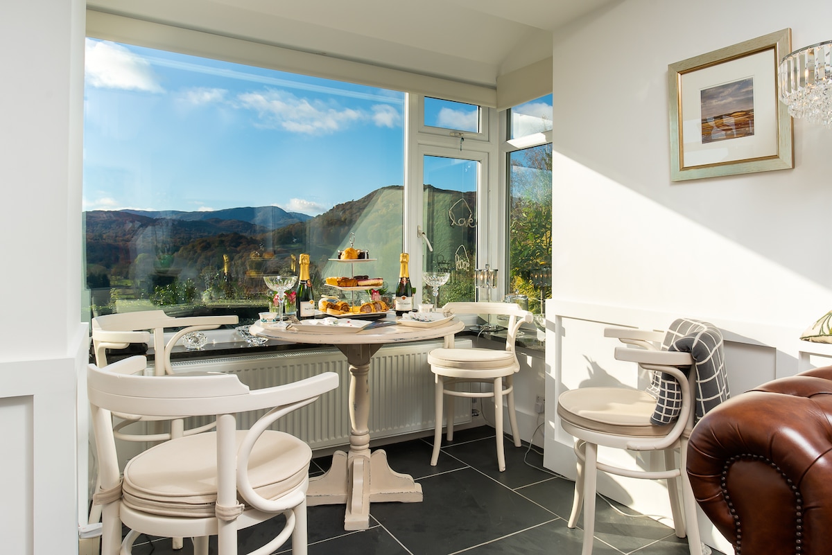 A dining area features a round table set with a tiered tray of treats, surrounded by four white chairs. Large windows provide expansive views of the surrounding hills, with sunlight illuminating the space. A framed art piece and a chandelier add elegant touches.