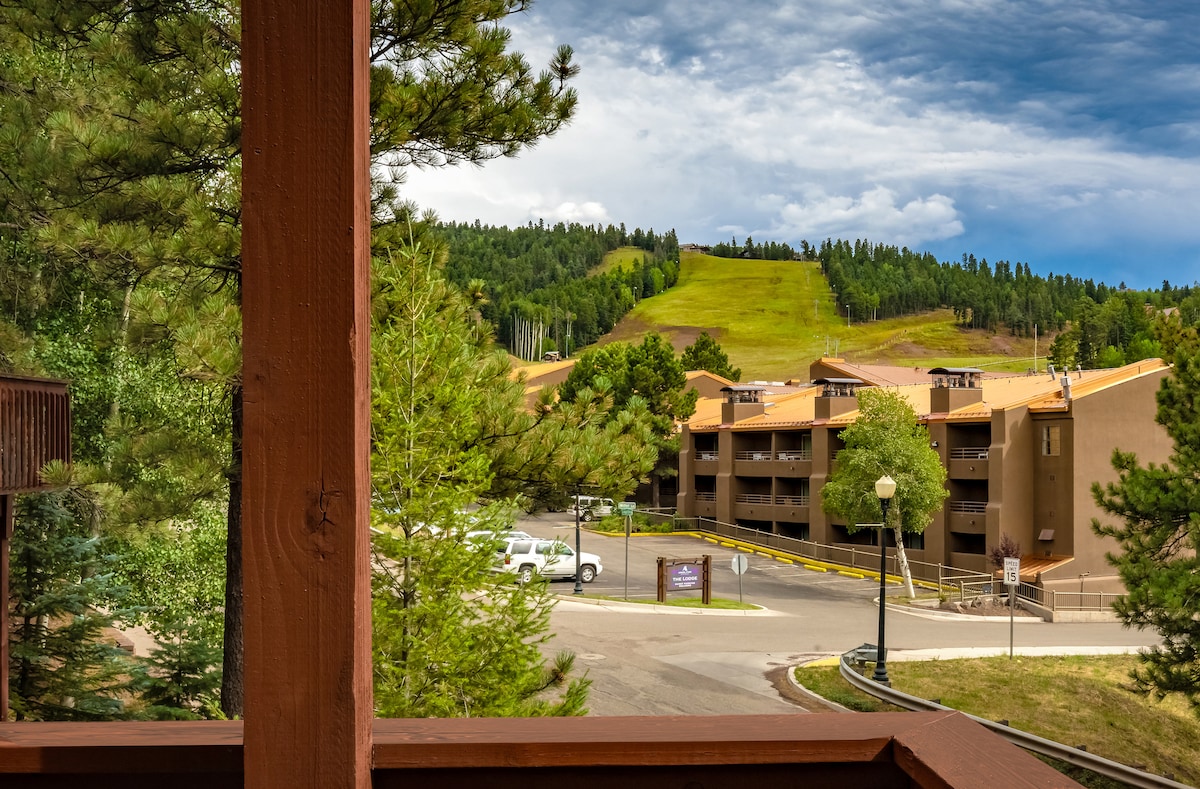 The view from the balcony showcases a landscape of rolling hills and lush greenery. A multi-story building is visible in the background, with vehicles parked nearby. The sky holds a mix of clouds and blue, enhancing the serene mountain setting.