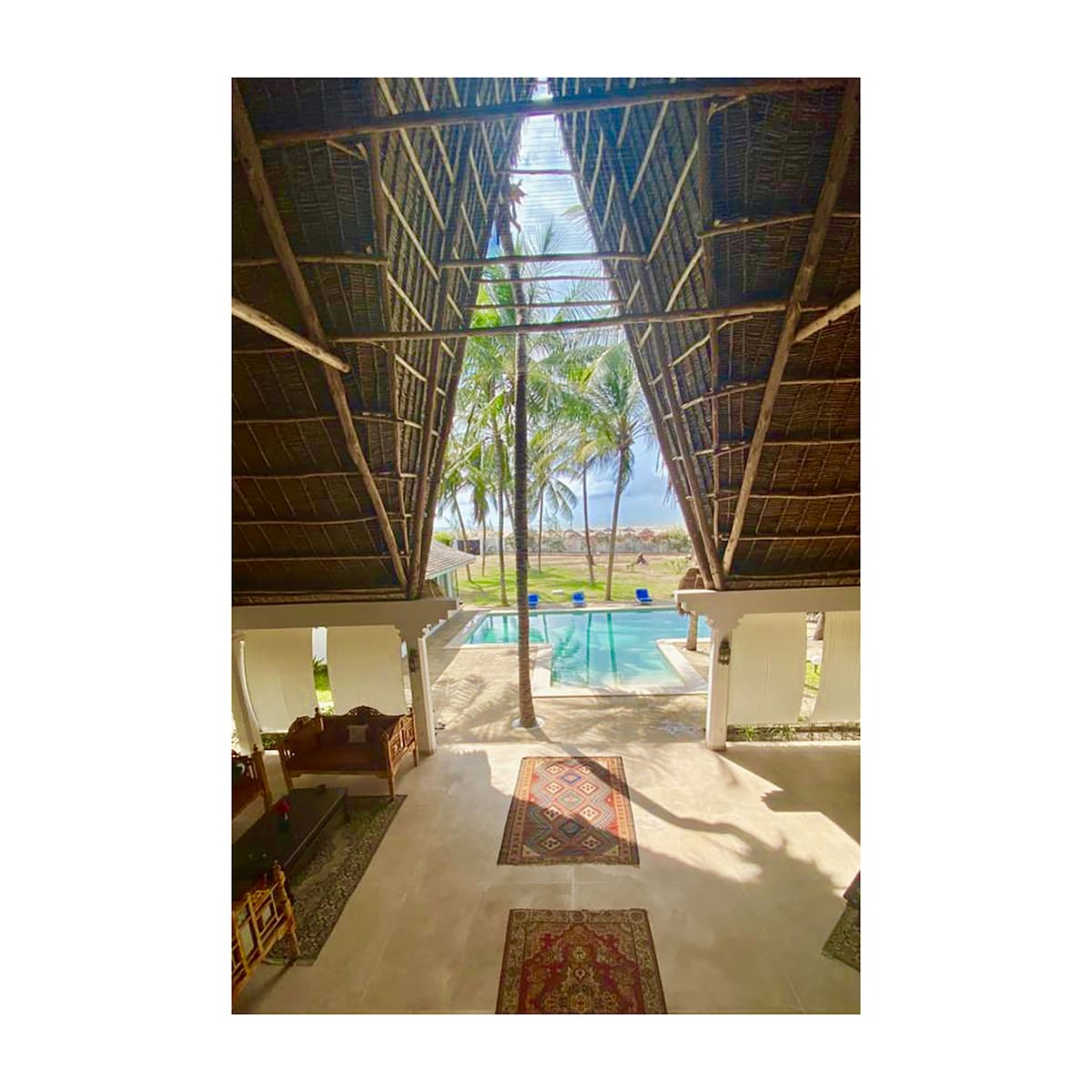 An architectural view displays a high thatched roof, framing a serene outdoor space with palm trees and a private pool. Elegant rugs are placed on the polished floor, leading towards an inviting seating area, with bright natural light streaming in from the open layout.