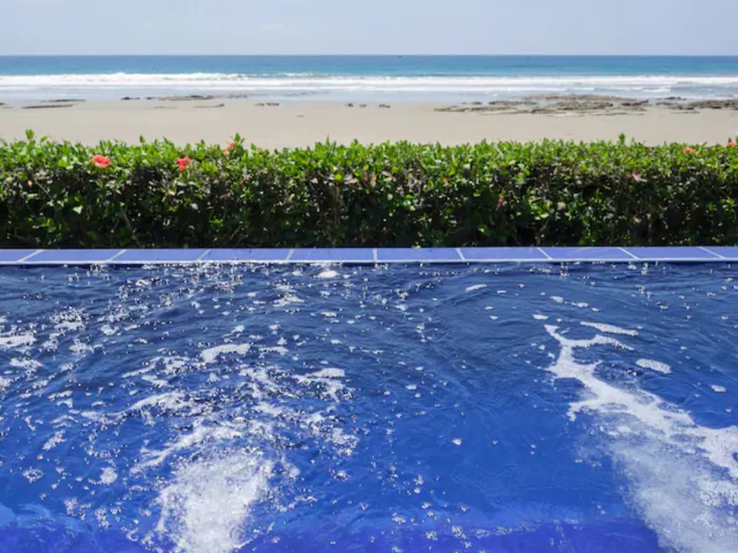 An infinity jacuzzi pool is displayed, with clear blue water and gentle bubbles reflecting sunlight. The edge of the pool seamlessly meets the sandy beach, lined with vibrant greenery. The ocean waves are visible in the distance, enhancing the serene coastal view.
