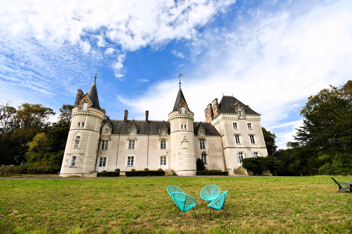 The exterior of a charming chateau is displayed, featuring two prominent turreted towers. A verdant lawn is in the foreground, accented by two teal patio chairs. The structure's stone façade is complemented by a backdrop of blue skies and scattered clouds.