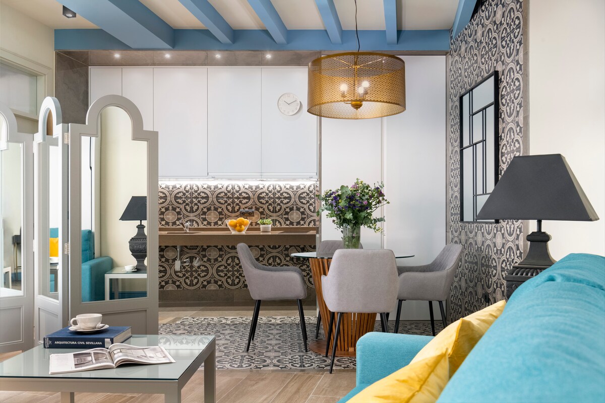 A bright and open living space is defined by a light blue ceiling and patterned accent wall. A dining table with gray upholstered chairs is complemented by a stylish lamp and floral centerpiece. A cozy blue sofa enhances the inviting atmosphere of the room.