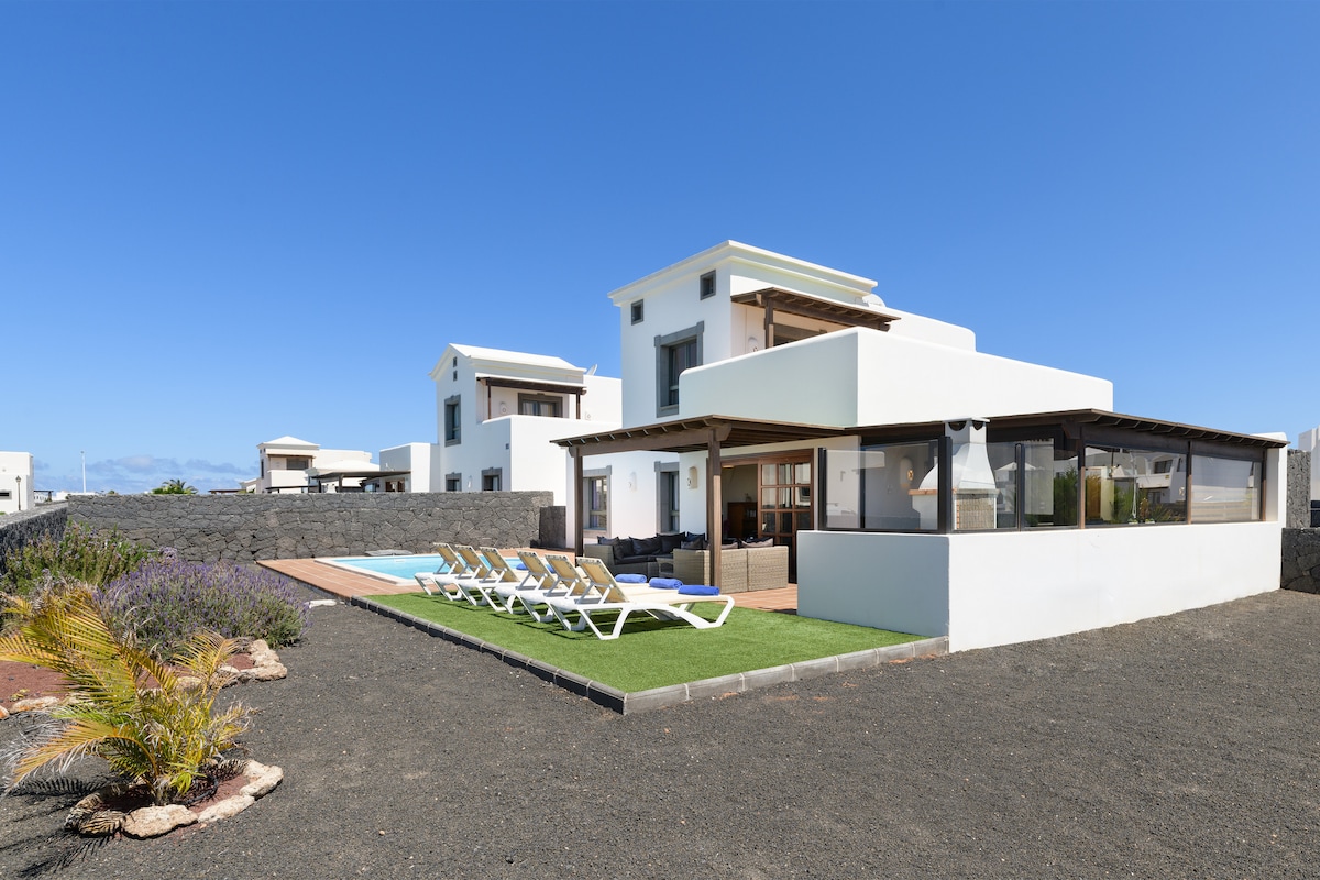 The villa is showcased surrounded by a well-maintained garden, featuring a private pool and sun loungers. A spacious terrace with outdoor seating is depicted, and neighboring structures are visible in the background under a clear blue sky.