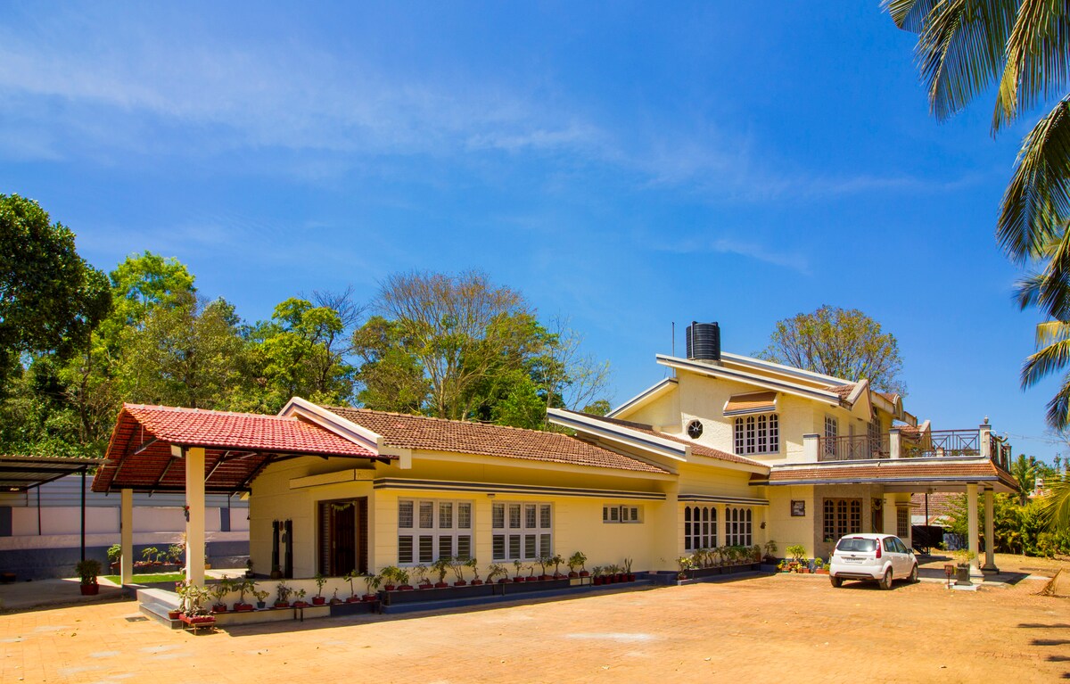 A charming homestay is presented, featuring a warm yellow exterior and a distinctive sloped red roof. The spacious front yard is lined with greenery, while a white vehicle is parked nearby. Clear blue skies and lush trees create a serene backdrop, enhancing the tranquil setting.