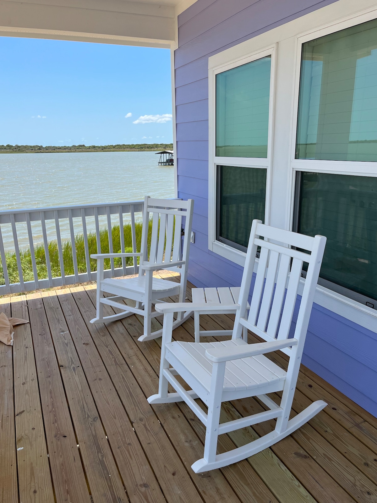 Two white rocking chairs are positioned on a wooden deck overlooking a calm body of water. Large windows provide a view of the outside, while the soft hue of the purple exterior complements the serene setting.