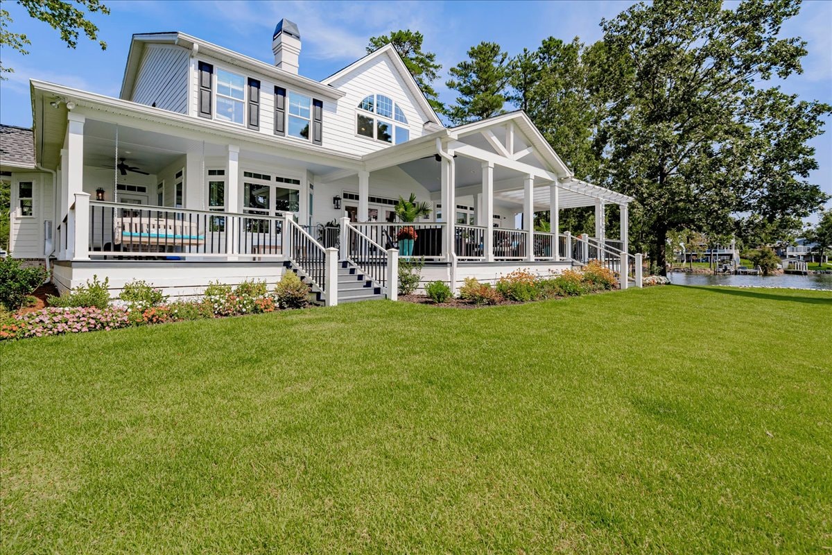 A charming lake house with a large front porch is surrounded by a well-maintained lawn. The exterior features white siding and a sloped roof, while nearby greenery creates a natural backdrop. Stairs lead down from the porch to the yard, providing easy access to the outdoor space.
