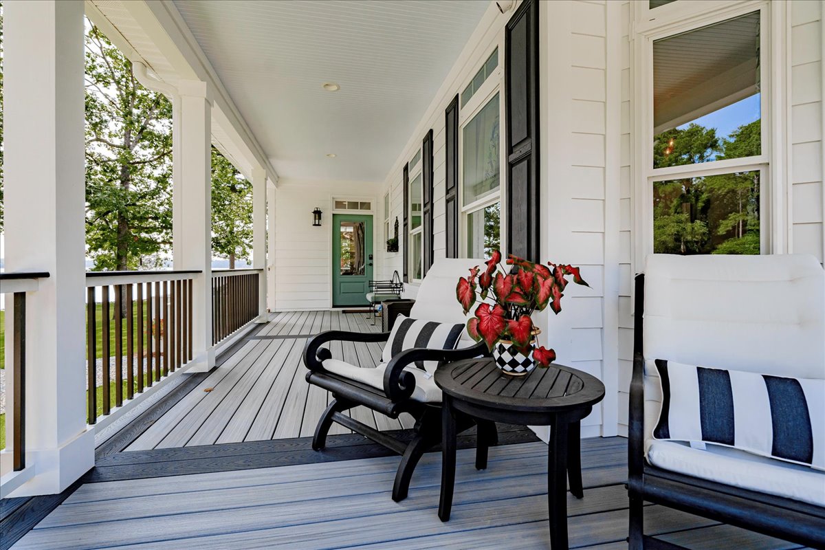 The covered porch features black and white striped seating arrangements, accompanied by a decorative table holding a vase of flowers. Large windows and a green door provide a welcoming entry, while the gray planked flooring extends toward the scenic views offered by the surrounding nature.