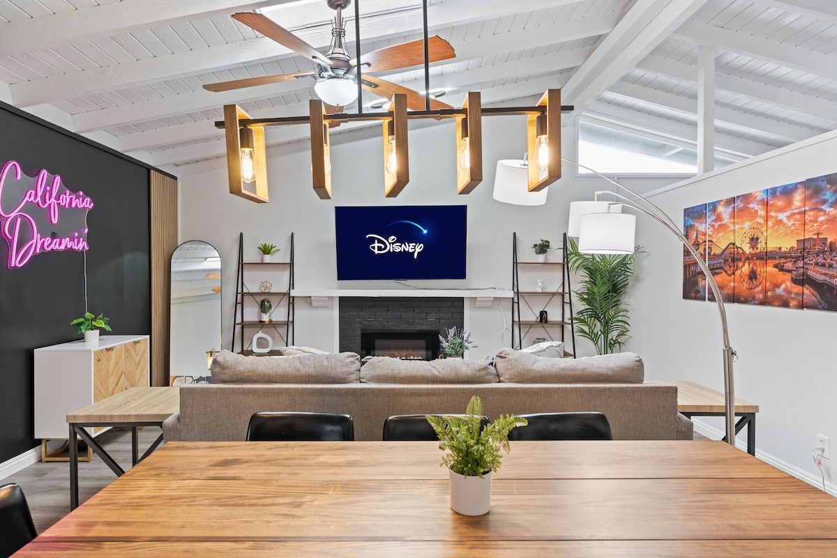 A spacious living area features a large sectional sofa facing a flat-screen TV. A dining table sits below a modern light fixture, surrounded by stylish decor. Warm wooden tones and soft greenery complement the open and inviting design, illuminated by natural light from the large windows.