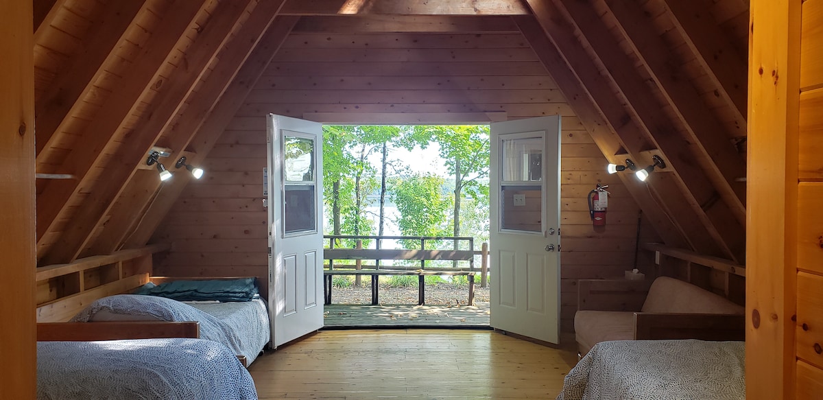 An open room features two double beds with natural light streaming in through large glass doors. The vaulted wooden ceiling adds warmth, while the view displays trees and outdoor seating amidst a peaceful landscape.