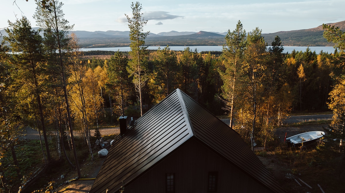 An aerial view captures the newly built house nestled among trees, showcasing a sloped roof. The surrounding landscape features a vibrant mix of autumn foliage, with a serene lake and distant mountains visible in the background under a clear sky.