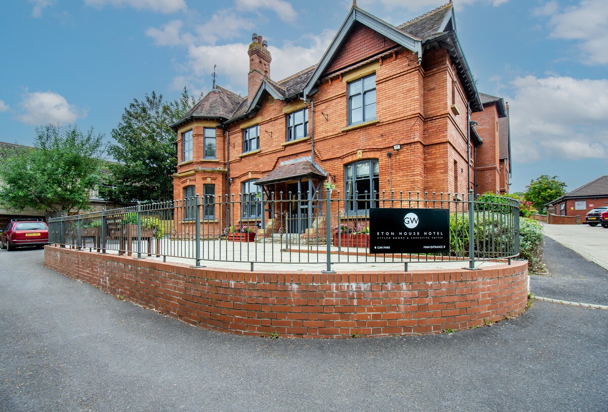 A charming three-story red brick building is surrounded by well-maintained greenery. A welcoming entrance with large windows is highlighted by a gated patio area. Ample parking space is visible in front, contributing to a sense of accessibility and inviting atmosphere.