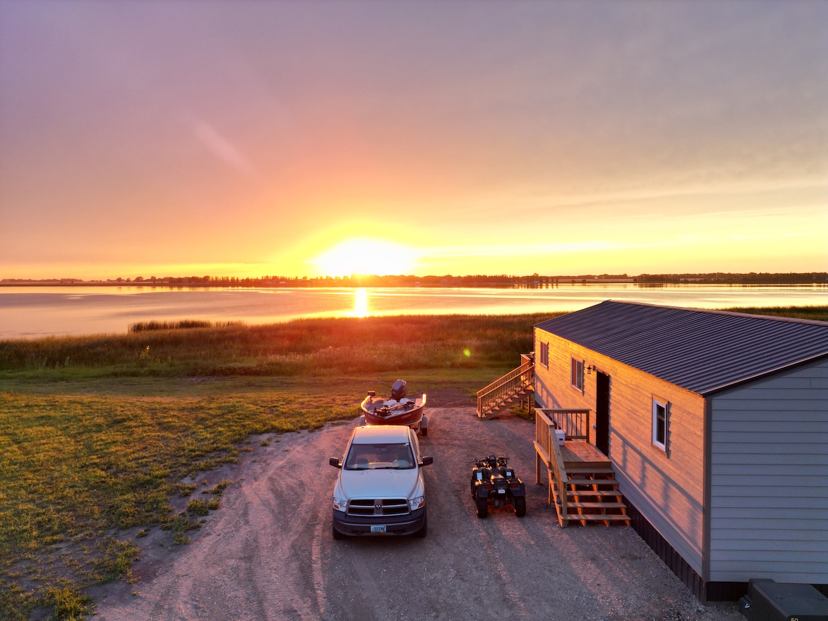The image captures a cabin positioned near a serene lake at sunset, with vibrant hues of orange and purple reflected in the water. A parked vehicle and an all-terrain vehicle are visible alongside the cabin, with grassy areas leading to the shore.