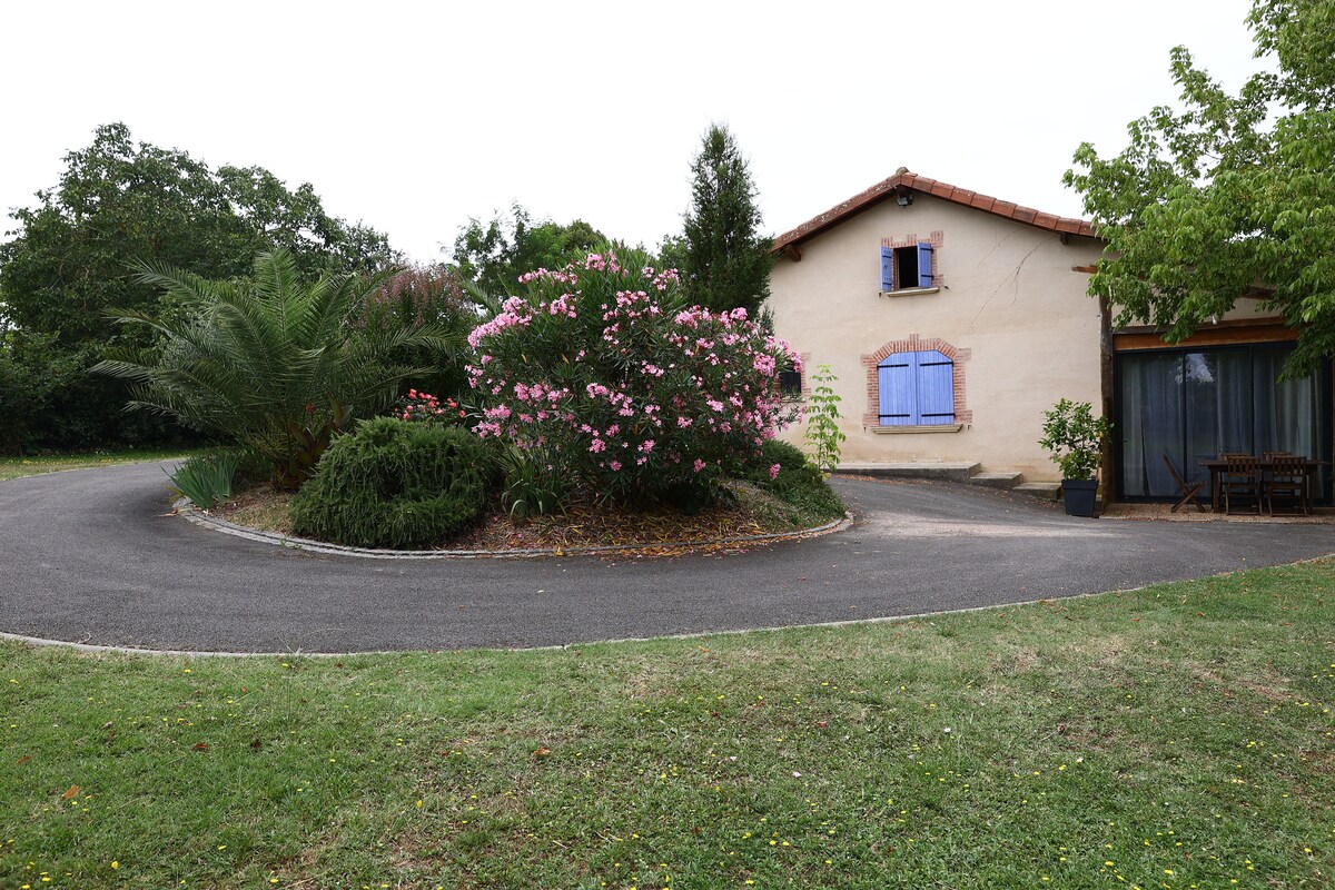 The exterior of the property is showcased with lush greenery and blooming flowers. A circular driveway leads to a charming house featuring purple-shuttered windows. A terrace is visible, providing outdoor seating amid a serene garden setting.