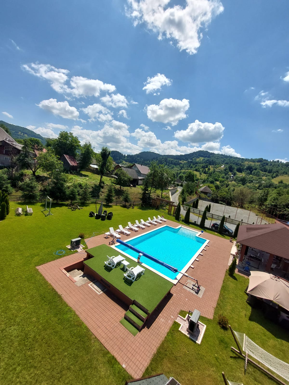 An outdoor swimming pool occupies the center of a well-kept green area, surrounded by lounge chairs. Lush trees and distant mountains provide a scenic backdrop under a bright blue sky filled with fluffy white clouds.