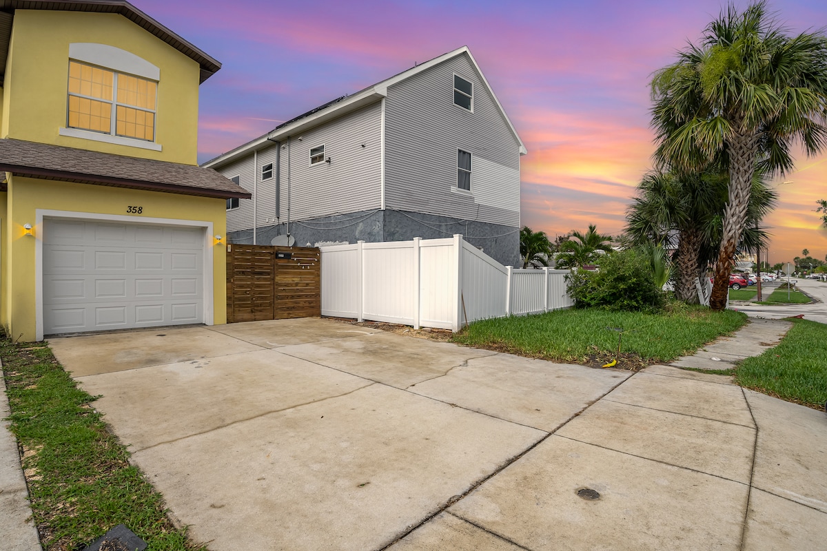 The image captures a residential driveway featuring a concrete surface leading to a garage. A fence provides privacy alongside the driveway, with grassy areas visible. A pastel sunset sky reflects soft colors above the neighboring house, enhancing the tranquil neighborhood setting.
