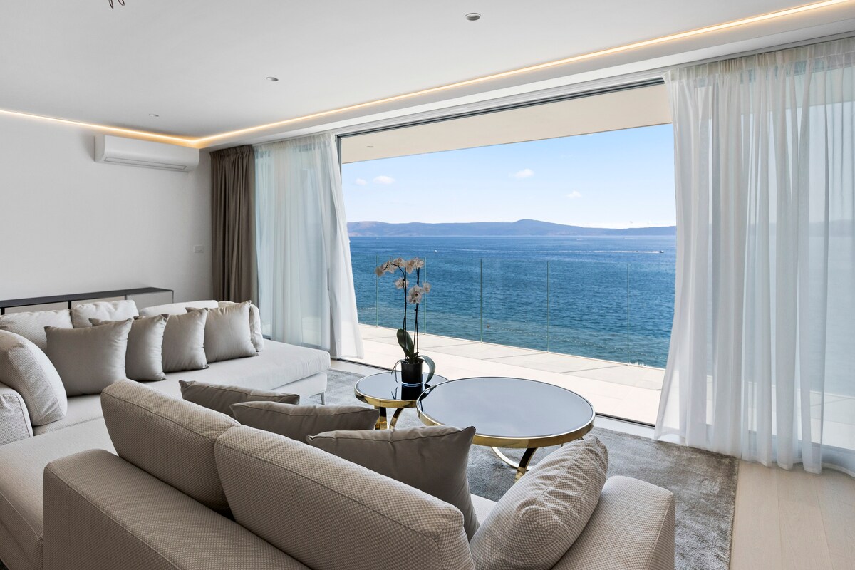 A spacious living area presents a large sectional sofa adorned with several cushions. A sleek coffee table sits in front, and expansive glass doors lead to a terrace with direct views of the sea, complemented by flowing sheer drapes that frame the scene.