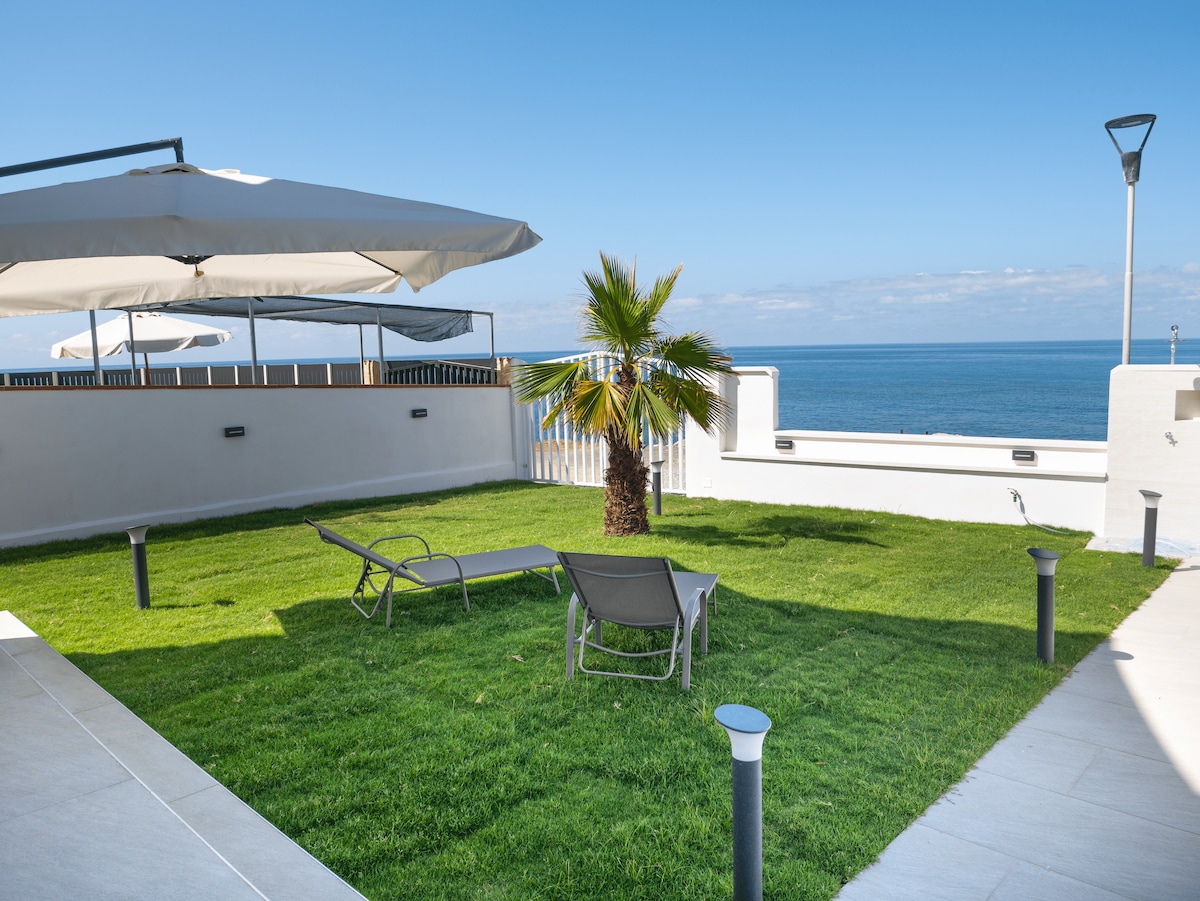 A private garden is presented with lush green grass, accentuated by a palm tree. Two sunbeds are positioned for relaxation. The sea can be seen in the background, complemented by clear blue skies and minimalistic lighting fixtures along the perimeter.