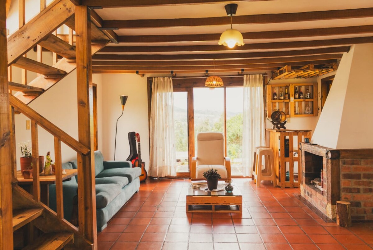 A comfortable living area features a light blue sofa and an armchair facing a large window, providing views of the surrounding nature. A wooden coffee table sits in front, with a simple decorative arrangement. A fireplace and wooden shelves enhance the rustic ambiance.