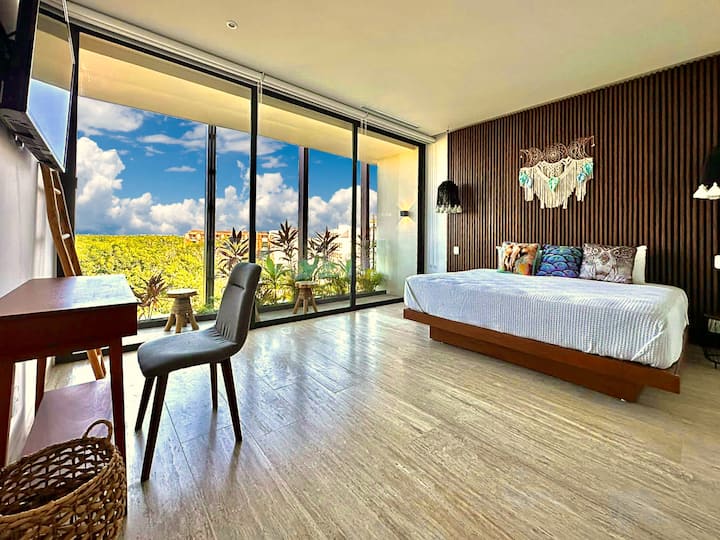 غرفة نوم رئيسية بسرير بمقاس كبير وتلفزيون ذكي كبير ومكتب عمل وتراس خارجي مع إطلالات جميلة على الغابة