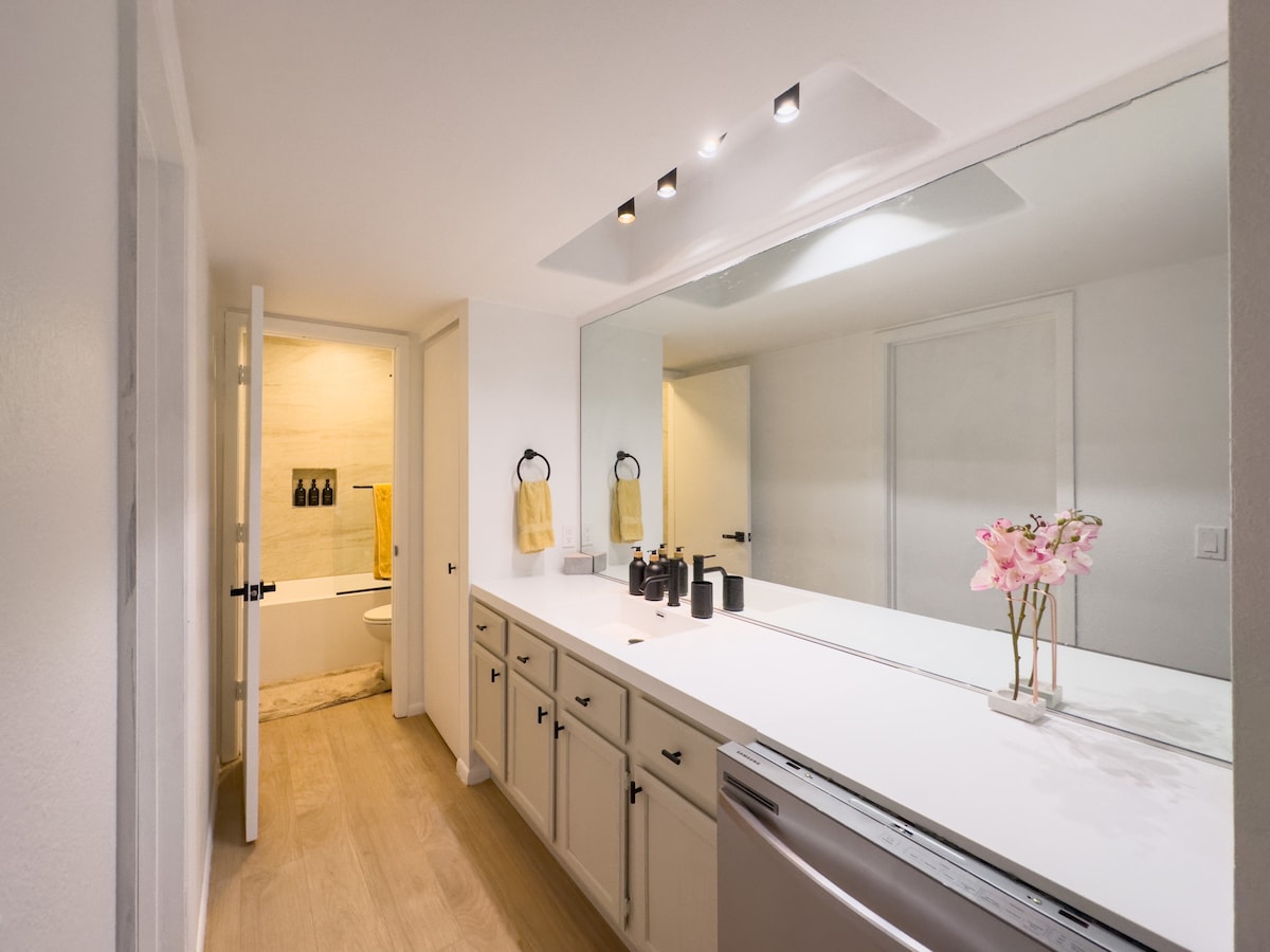 A bright bathroom is featured with a wide, white countertop and dual sinks. Below, cabinets provide storage. A large mirror reflects the light fixtures above. A tasteful arrangement of towels and a small vase with flowers are positioned near the sinks, enhancing the simple elegance of the space.
