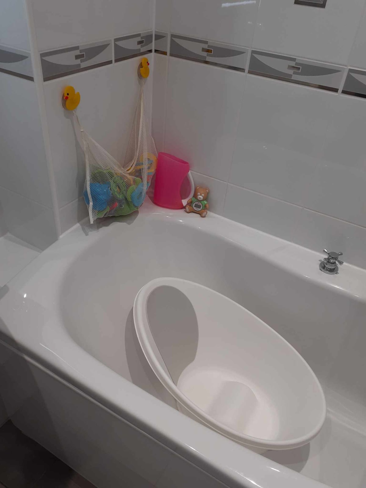 A clean bathroom features a curved bathtub filled with natural light. A white baby bath is positioned inside, alongside a colorful mesh bag containing bath toys. Pink and decorative items are displayed on the surrounding surfaces, adding a touch of charm to the space.