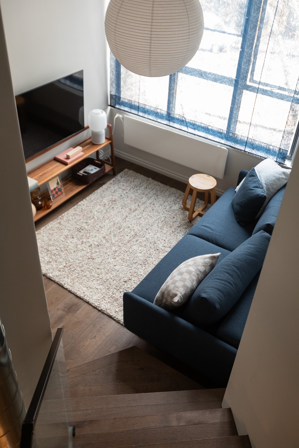 The living area features a dark sofa with plush pillows, positioned adjacent to a light-colored area rug. A wooden side table and a low TV cabinet are visible, along with a large window dressed in sheer blue curtains. Light filters softly into the space.