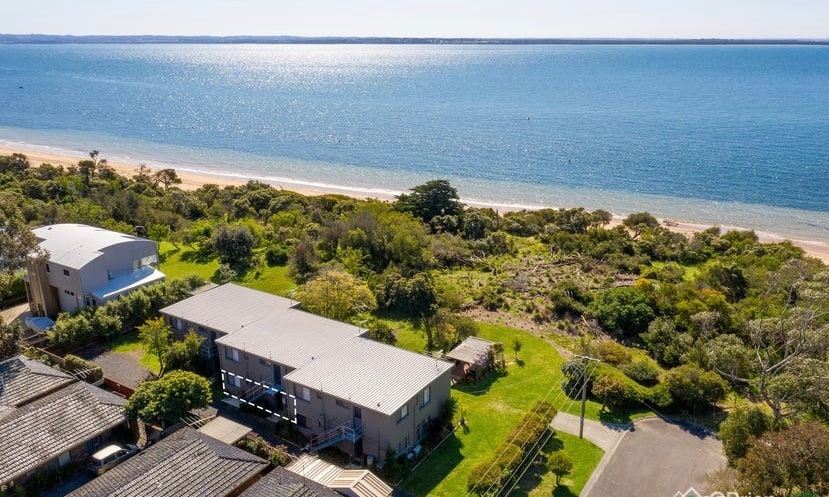 An aerial view showcases the boutique complex as it sits adjacent to a peaceful beach. Lush greenery surrounds the area, with the calm water reflecting the sunlight. Nearby buildings are visible, along with sandy expanses that lead to the shoreline.
