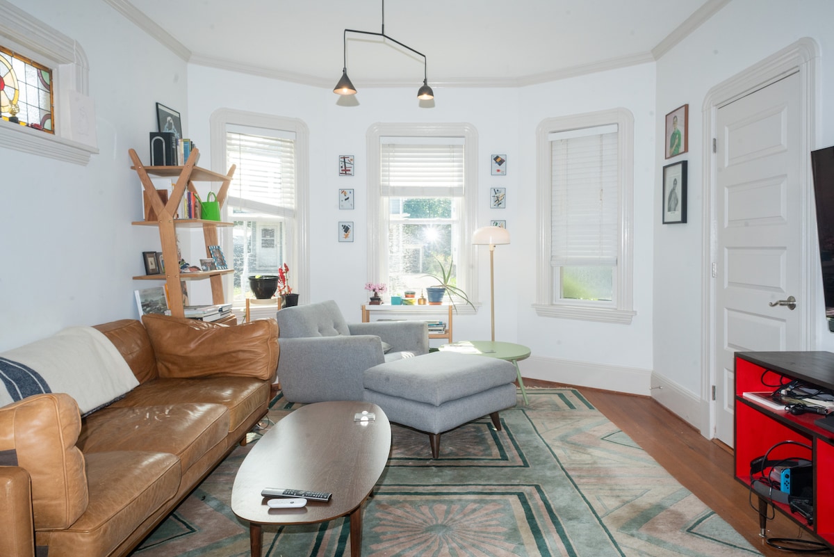A spacious living room is highlighted by a large, light brown leather sofa and a sleek gray armchair. Natural light enters through multiple windows, illuminating a circular area rug. A wooden coffee table and a modern light fixture add to the inviting atmosphere.