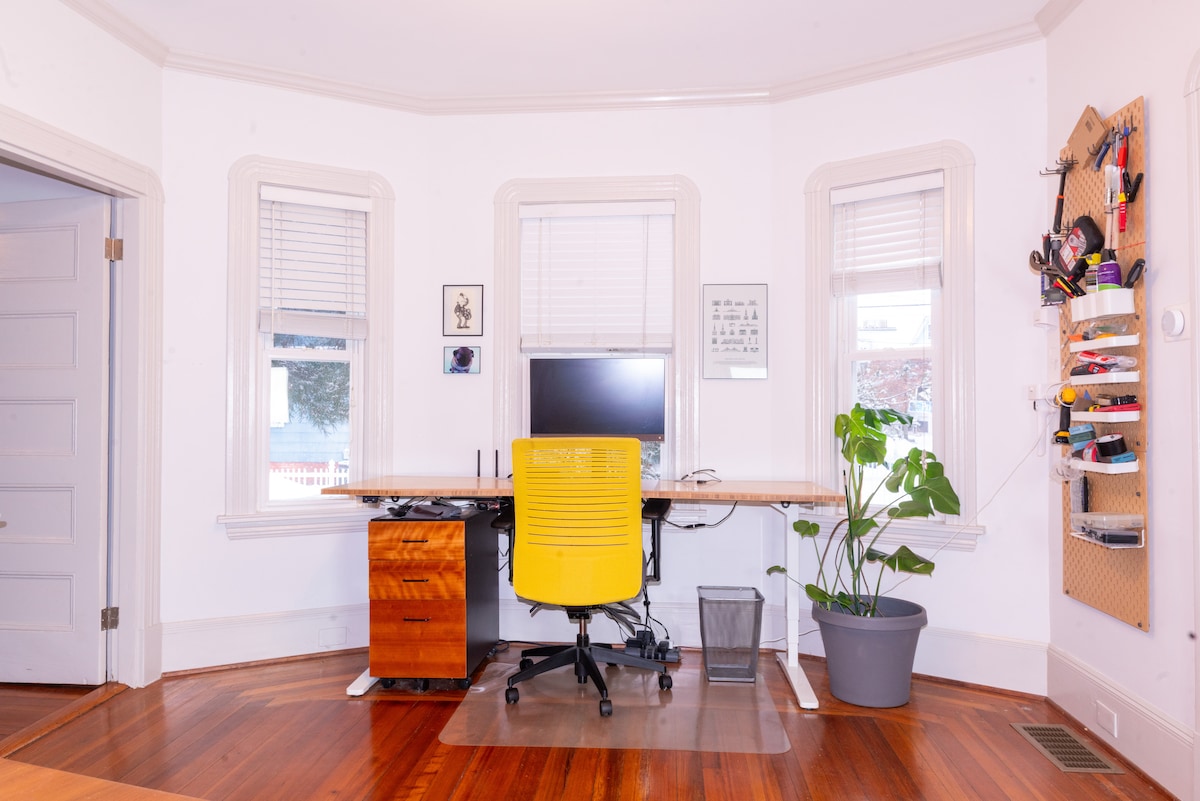 A dedicated workspace is presented with a large wooden desk and a yellow office chair. A monitor is positioned on the desk, while natural light filters through three windows with blinds. A potted plant occupies a corner, and various tools are neatly organized on a wall-mounted pegboard.