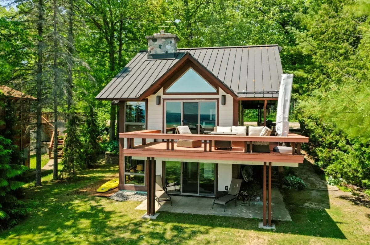 A modern two-story house is nestled among lush greenery, featuring large windows that provide an abundance of natural light. A spacious deck is visible, furnished with outdoor seating and a parasol. The surrounding landscape incorporates trees and a well-maintained lawn.