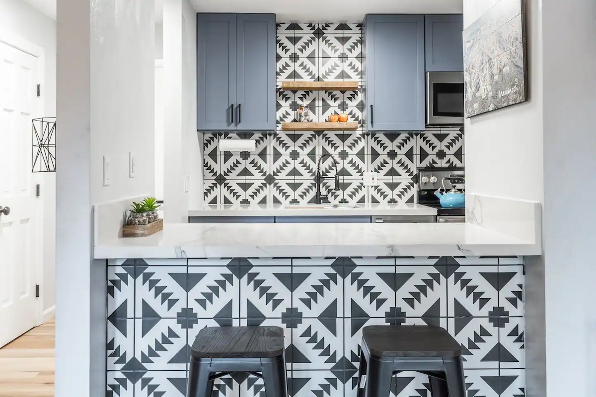 A kitchen area features modern cabinetry in deep blue, highlighted by a striking patterned backsplash in black and white. Open shelving displays decorative items, and a microwave is visible. Two black stools are positioned at a counter topped with white quartz.