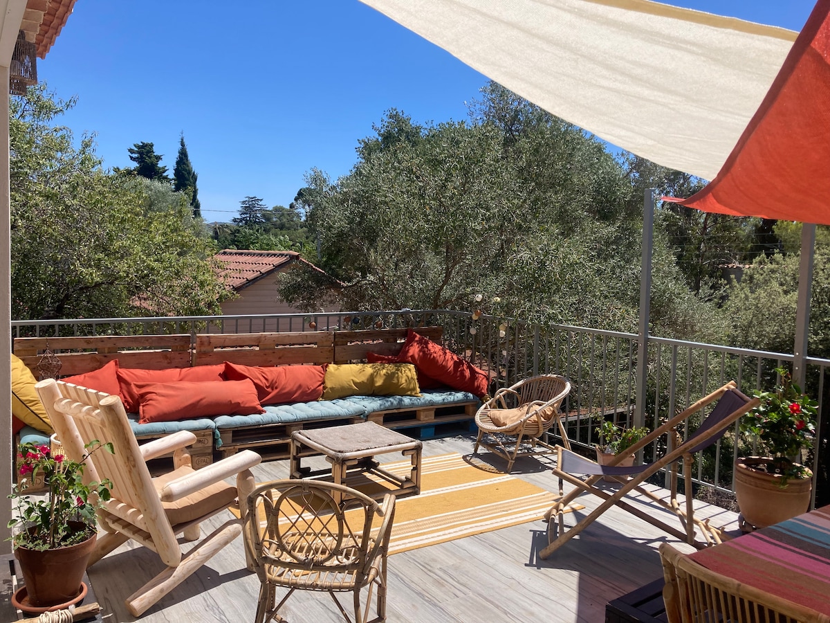 A sunlit terrace is furnished with a long, cushioned seating area adorned with colorful pillows. A small table sits in the center, accompanied by wooden chairs. Shade is provided by fabric canopies, creating a comfortable outdoor space surrounded by greenery.