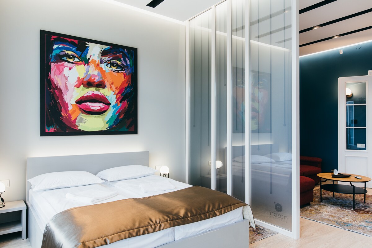 The bedroom features a spacious bed with a light duvet, surrounded by softly glowing bedside lamps. A glass partition separates the sleeping area from the lounging space, which includes a cozy sofa and a stylish wall art piece, enhancing the room's modern decor.