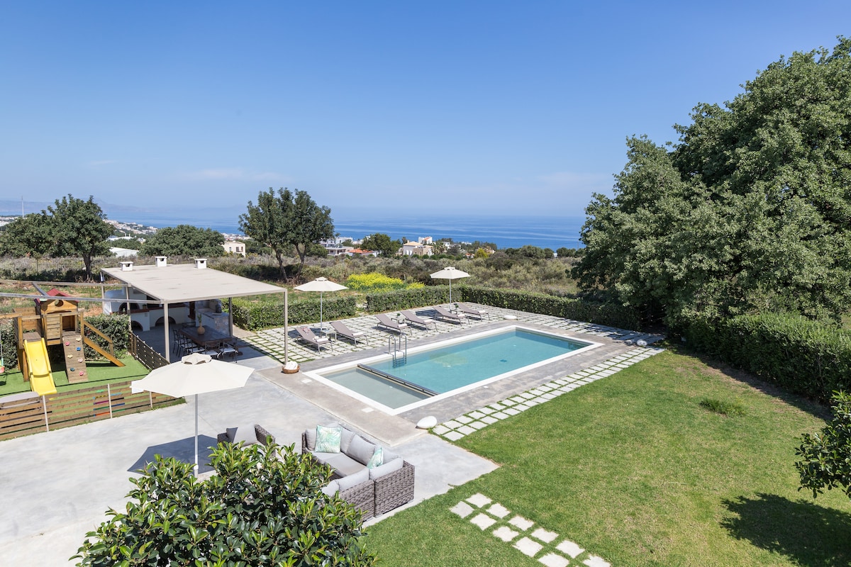 An outdoor area features a spacious private pool surrounded by sunbeds and umbrellas. Nearby, a children’s playground with slides is partially visible. Lush greenery and views of the Cretan sea create an inviting atmosphere for relaxation and family fun.