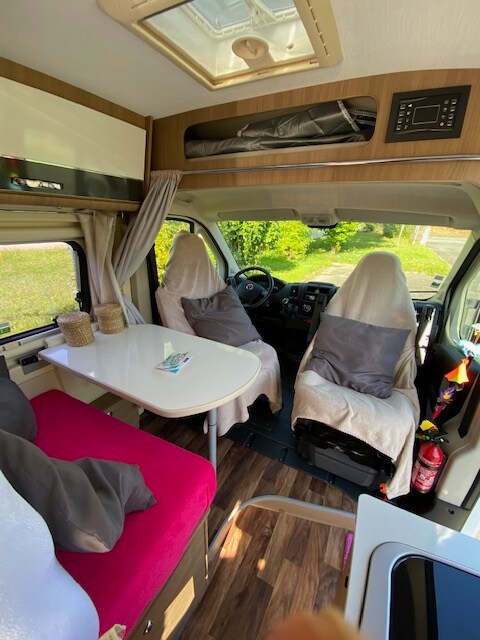 The interior features a functional dining area with a table set against a warm-toned backdrop. Two comfortable seats, covered with light fabric, are positioned in the front. Natural light enters through the overhead window, enhancing the cozy environment.