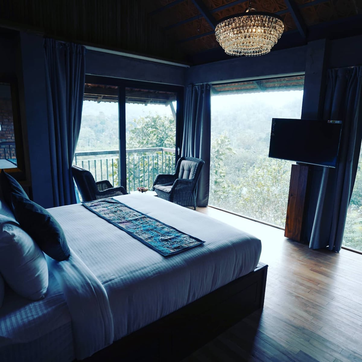 A spacious bedroom features a large bed with a decorative runner, flanked by two wicker chairs. Floor-to-ceiling windows provide a scenic view of the surrounding greenery, while a chandelier adds a touch of elegance to the warm wooden accents.