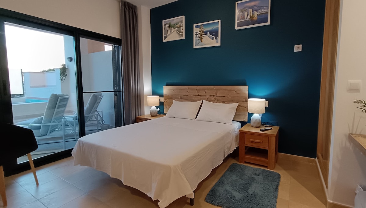 A bright bedroom is featured with a spacious bed and nightstands on each side. Soft lighting is provided by bedside lamps. The wall behind the bed is painted in a deep blue, while photographs add a decorative touch. Large glass doors offer views of the terrace and pool area.