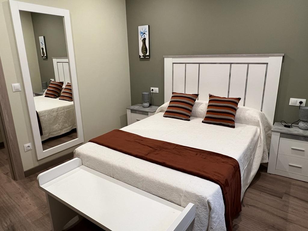 The room features a spacious bed adorned with patterned cushions and a coordinating throw. A large mirror reflects the bed area, enhancing the sense of space. Nightstands with lamps are positioned on either side of the bed, while muted green walls create a calm environment.