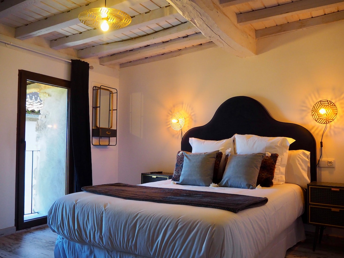 A spacious sleeping area features a large bed with neatly arranged cushions, complemented by warm wall sconces providing gentle illumination. Natural light filters in through a glass door, which opens to a balcony, enhancing the airy feel of the room's design and wooden ceiling beams.