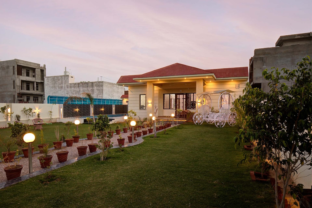 A charming villa is surrounded by a lush garden featuring neatly arranged flower pots and soft lighting. A well-maintained lawn provides ample space for outdoor activities, while elegant seating arrangements are visible. The entrance showcases large glass doors, inviting nature inside.