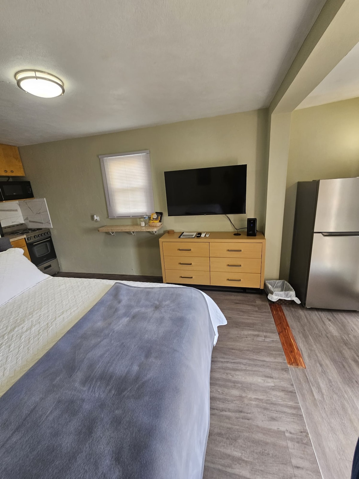 A studio space features a spacious bed covered with a light-colored quilt. A wooden dresser with multiple drawers stands opposite the bed, accompanied by a wall-mounted television. The kitchenette is visible, showcasing appliances and cabinetry, while a sleek refrigerator occupies a corner.