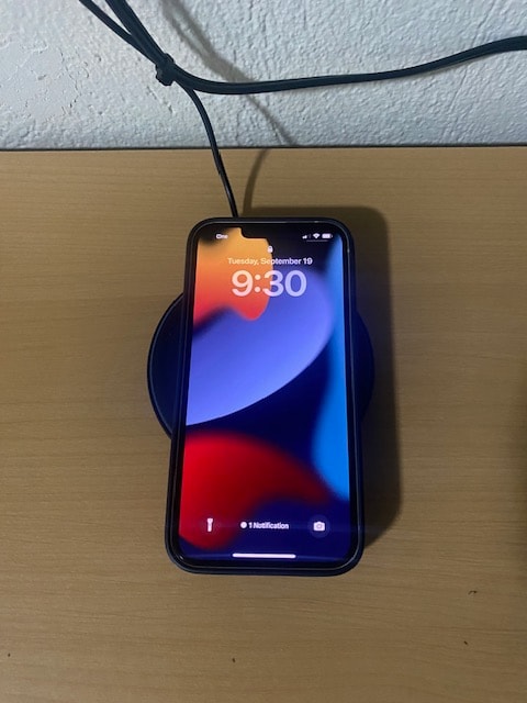 A smartphone is seen resting on a wireless charging pad, displaying the time as 9:30. The background features a wooden surface with a neutral tone, and a cable is plugged into the device, suggesting connectivity.