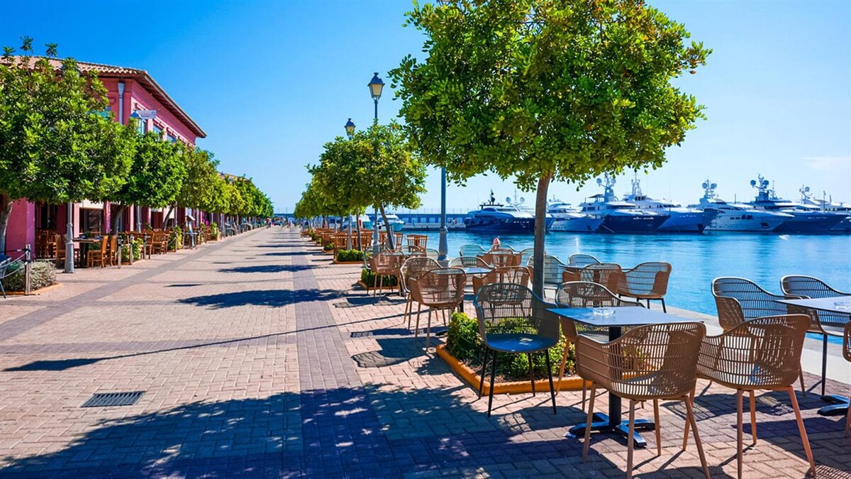 A waterfront promenade is lined with evenly spaced trees and outdoor seating. Wicker chairs and glass tables provide a relaxed dining option. The sea glimmers in the background, with yachts moored along the dock, creating a calm coastal atmosphere.
