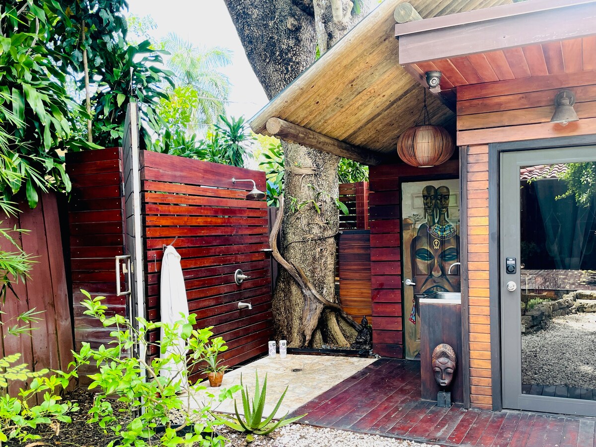 An outdoor shower area is framed by warm wooden walls and lush greenery. A natural stone floor complements the wooden deck. A large tree stands prominently nearby, enhancing the tropical ambiance. The shower features a modern fixture, and personal items can be seen resting on the ground.