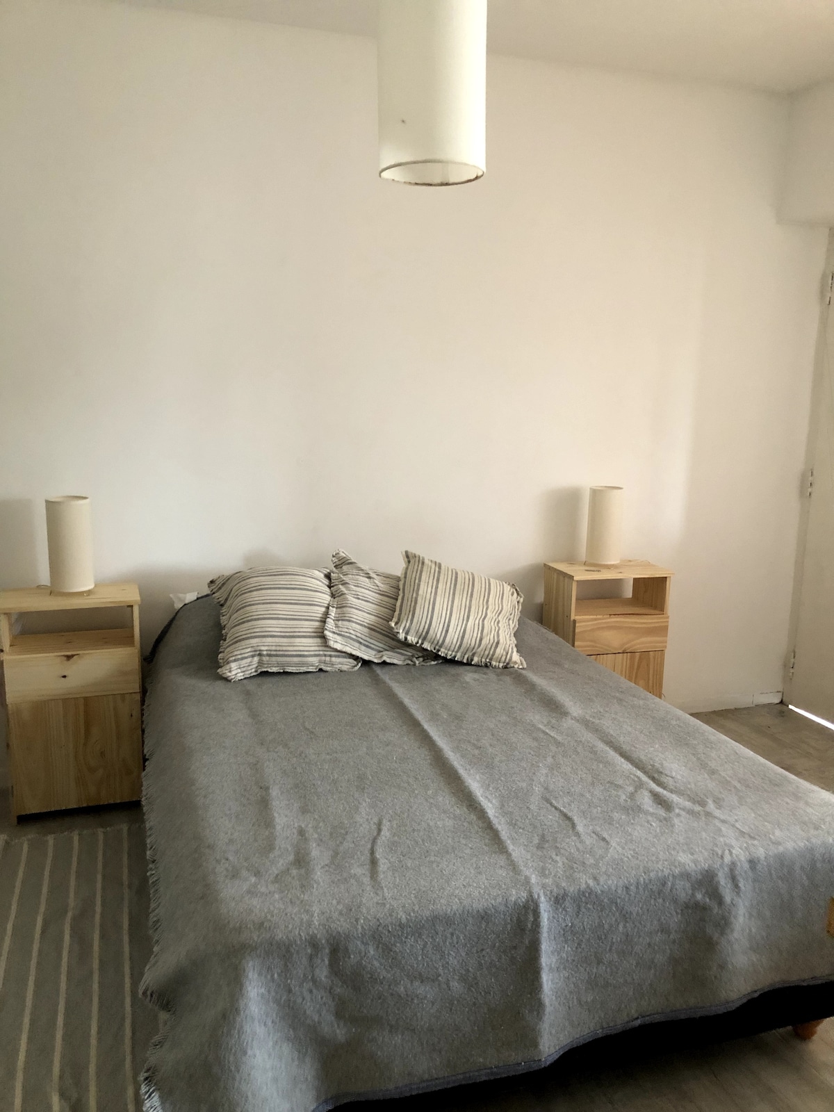 A serene sleeping area is present, featuring a spacious bed dressed in a simple grey blanket. Two wooden nightstands are positioned on either side, each topped with a modern lamp. Soft natural light is visible streaming in from a nearby window.