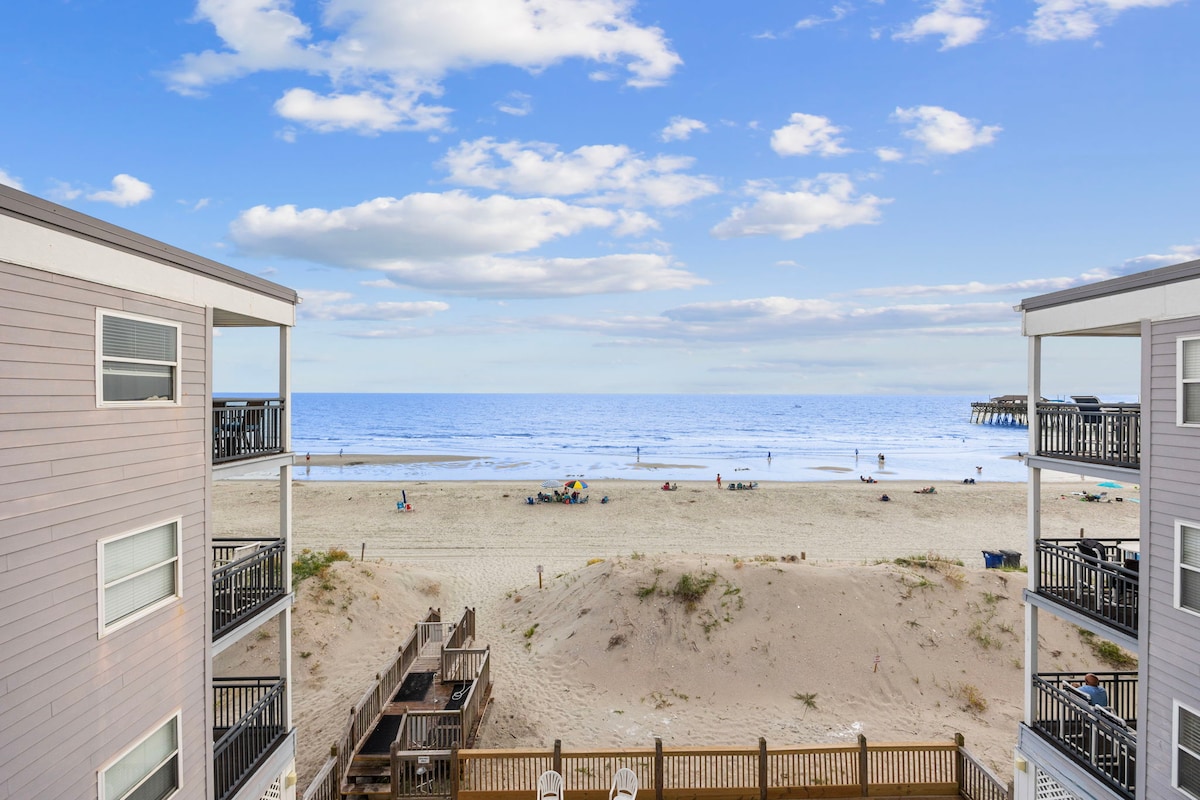A clear view of the beach and ocean is presented from a balcony between two buildings. Soft sand leads to the gently lapping waves, with people enjoying the shoreline and the iconic pier visible in the distance under a partly cloudy sky.